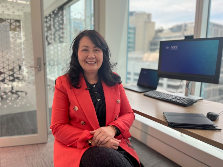 A woman with mid length brown hair sits looking into camera and smiling. She's wearing a bright red jacket and has her hands clasped in front of her.
