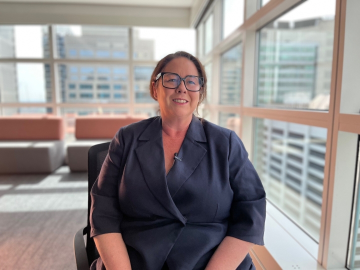 A woman with her hair tied back looking direct to camera and smiling. She is in an office environment and is wearing a navy top.